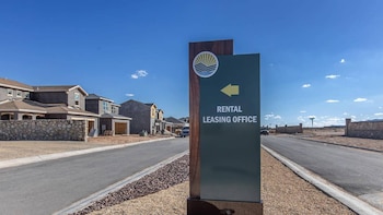 Un cartel verde oscuro con texto blanco 'RENTAL LEASING OFFICE' y una flecha amarilla a la izquierda, junto a una carretera pavimentada y casas en construcción al fondo