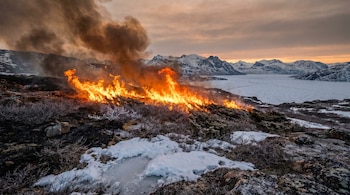 Incendio con llamas intensas y humo, en un terreno con arbustos y nieve. Al fondo, montañas nevadas y un lago congelado bajo cielo nublado.