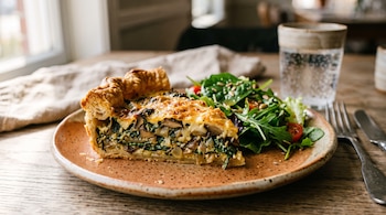Una porción de tarta horneada con ensalada verde en un plato de cerámica sobre una mesa de madera con un vaso de agua.