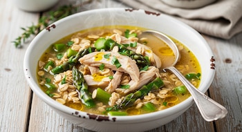 Tazón blanco con sopa caliente de avena, trozos de pollo y espárragos verdes. Una cuchara de metal está en el tazón. Hay vapor y hierbas picadas.