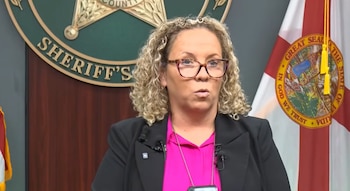 Mujer con cabello rizado y gafas, vistiendo una camisa rosa y un blazer negro, habla frente a un emblema de la oficina del sheriff y la bandera de Florida