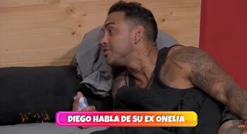 Diego Chávarri, un hombre con tatuajes en brazos y cuello, recostado en la cama sobre almohadas grises, con una pared de madera y una roja al fondo