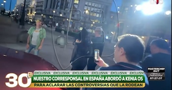 Una calle al atardecer, con un reportero sosteniendo un teléfono frente a un vehículo oscuro. Una persona con camisa verde y jeans rotos se ve a la izquierda