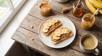 Dos tostadas con crema de cacahuete y plátano en un plato, café, tarros de crema de cacahuete, miel y un racimo de plátanos sobre una mesa de madera