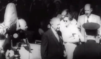 Imagen en blanco y negro de Hendrik Frensch Verwoerd, un hombre mayor con traje, caminando entre varias personas y una cámara de cine antigua