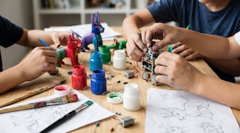 Varias manos arman un robot con piezas electrónicas y pintan figuras de alebrijes. En la mesa hay botes de pintura acrílica, pinceles y bocetos.