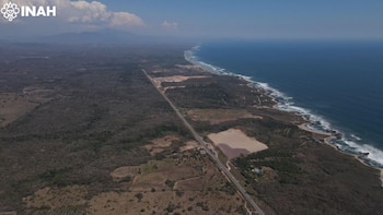 Fotografía aérea de una carretera que bordea la costa, con mar azul y olas, vegetación seca, montañas al fondo y el logo del INAH