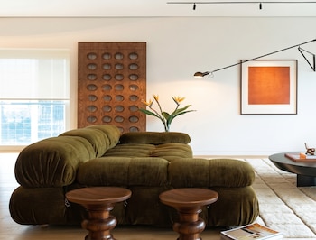 Salón moderno con sofá modular verde oliva, taburetes de madera, mesa de centro negra, alfombra clara, panel de madera decorativo y cuadro naranja en la pared.