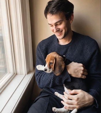 Un hombre joven con suéter de color azul oscuro sostiene un cachorro de raza beagle tricolor. Ventana con marco blanco a la izquierda