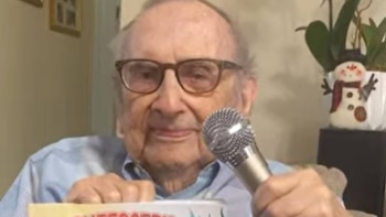 Un hombre mayor con gafas y camisa azul sostiene un micrófono con una mano y un objeto blanco con la otra. Un muñeco de nieve decorativo se ve al fondo