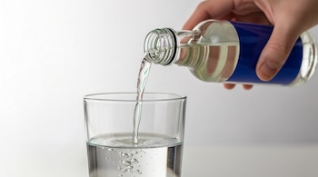 Primer plano de una mano que vierte un líquido claro desde una botella de vidrio con etiqueta azul en un vaso transparente lleno parcialmente de agua.