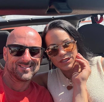 Un hombre calvo con barba corta y una mujer de cabello negro, ambos con gafas de sol, sentados dentro de un vehículo descapotable