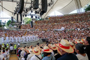 Escenario con once bailarines en trajes típicos, una bailarina de blanco al centro. Detrás, una orquesta. Al fondo, árboles y montañas. Piñas en el borde del escenario