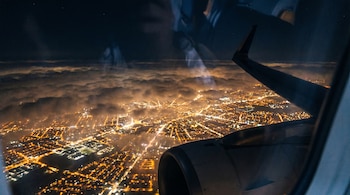 Vista nocturna desde la ventanilla de un avión: una ciudad iluminada con luces amarillas, nubes sobre ella, y el ala y motor del avión en primer plano.