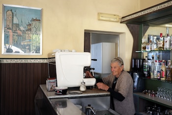 Mujer anciana con cabello gris y cárdigan marrón opera una máquina de café espresso blanca en un mostrador, con botellas, vasos y un cuadro en la pared