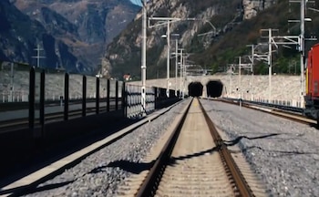 Vías de tren paralelas que conducen a dos túneles oscuros en un paisaje montañoso. Postes de catenaria y un tren rojo son visibles a la derecha
