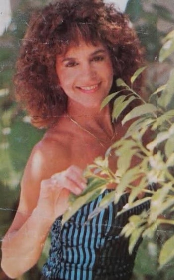 Retrato de Lady M sonriendo, con cabello rizado castaño, collar dorado y blusa a rayas azules y negras, detrás de hojas verdes