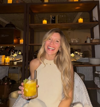 Laurita Fernández, de cabello rubio, sonríe ampliamente mientras sostiene un cóctel naranja en un bar ambientado con velas y estantes de madera