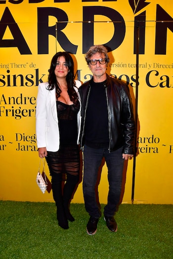 Una mujer con chaqueta blanca y un hombre con chaqueta de cuero negra posan frente a un muro amarillo con texto y suelo de césped artificial