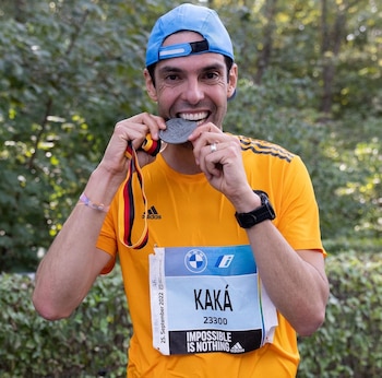Un hombre sonríe mordiendo una medalla con cinta de tres colores, lleva una gorra azul, camiseta naranja, dorsal con "KAKÁ" y reloj en la muñeca