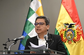 Hombre con gafas y traje oscuro habla en un podio con dos micrófonos. Detrás, se ven la Wiphala y la bandera tricolor de Bolivia