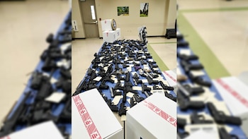 Decenas de pistolas y fusiles etiquetados sobre mesas con manteles azules y cajas de cartón selladas con cinta de seguridad