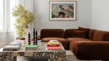 Vista interior de una sala de estar con sofá modular marrón, mesa de centro de mármol, libros, planta verde y una obra de arte colgada en la pared blanca.