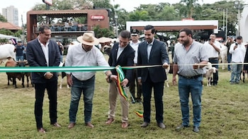 Cinco hombres, uno cortando una cinta tricolor, en un campo de hierba con vacas y cabras. Edificios y árboles al fondo bajo un cielo nublado