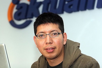 Un hombre con gafas, barba incipiente y sudadera con capucha marrón. Detrás de él, un logo azul y naranja borroso y una pantalla de laptop plateada