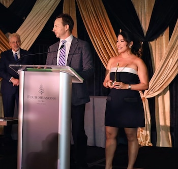 Un hombre en traje habla en un podio con el logo Four Seasons Houston. A su lado, una mujer sonríe y sostiene un trofeo dorado. Otro hombre está detrás