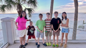 Seis niños, con sus rostros pixelados, de pie en un balcón con barandilla blanca, frente a un cuerpo de agua y palmeras.