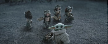 Grogu, un pequeño ser verde con orejas grandes, tira de un carrito sobre tierra oscura, con cuatro criaturas pequeñas con vestimentas de corteza detrás