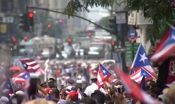 Multitud de personas ondeando banderas de Puerto Rico en un desfile urbano. Edificios y semáforos visibles de fondo, parcialmente desenfocados