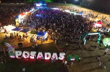 Vista aérea de un gran evento nocturno al aire libre con una multitud, un escenario iluminado, varios puestos y un letrero grande que dice "POSADAS"