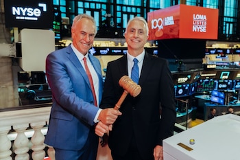 Dos hombres sonríen y sostienen un mazo de madera en un edificio bursátil. Pantallas digitales con el logotipo NYSE y un cartel rojo de Loma Negra se ven de fondo
