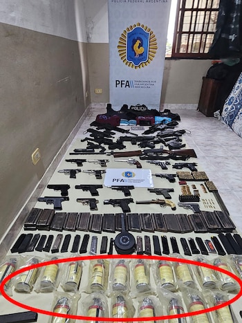 Decenas de pistolas, fusiles, cargadores, cajas de municiones y granadas de gas dispuestas en el suelo frente a un cartel de la Policía Federal