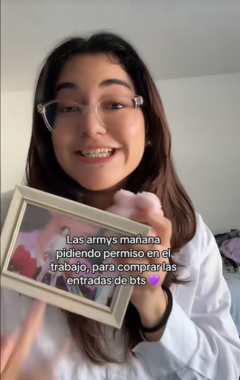 Mujer joven de cabello oscuro y gafas sonríe mientras sostiene una pequeña foto enmarcada de BTS, con un objeto rosa mullido cerca de su mano