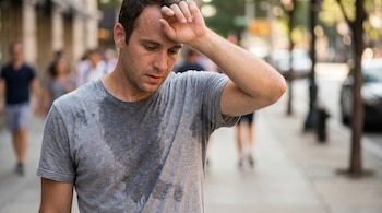 Primer plano de un hombre con remera gris sudando profusamente en una vereda, una gota cae por su cara mientras se limpia la frente, el fondo está difuminado.