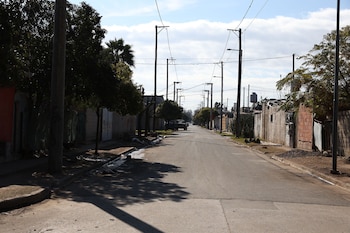 Vista de una calle de asfalto en un barrio residencial, flanqueada por casas bajas con cercos y árboles. Numerosos postes de servicios cruzan la vía bajo un cielo con nubes