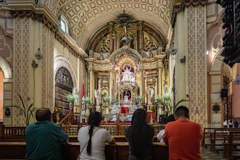 Vista trasera de varias personas arrodilladas en bancos de madera dentro de una iglesia con un altar dorado y ornamentado al fondo