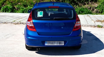 Parte posterior de un coche azul con un distintivo verde de la letra L en el cristal trasero y la matrícula difuminada.