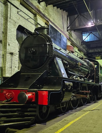 Locomotora de vapor negra con parachoques rojo, situada en un depósito con paredes de ladrillo y techo de estructura metálica