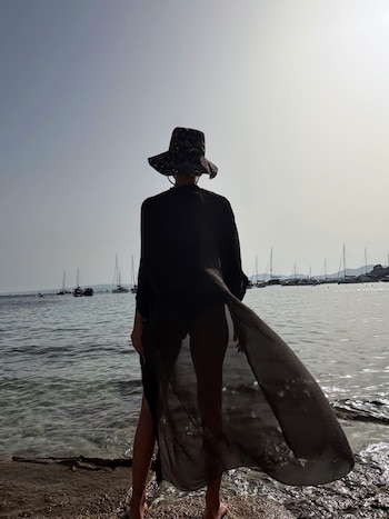 Una mujer de espaldas con sombrero y vestido playero oscuro en la orilla del mar. Barcos, veleros y botes se observan en el agua bajo un cielo claro