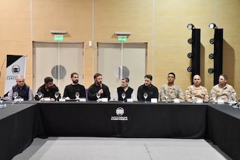 Nueve hombres vestidos de civil y con uniformes militares se encuentran sentados a una larga mesa con mantel negro
