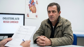 Hombre sentado frente a un médico que sostiene un informe. En pantalla se lee "Colesterol alto, Riesgo cardíaco". Un póster de corazón en la pared.