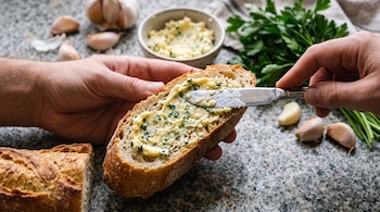 Manos que usan un cuchillo para untar mantequilla con ajo y perejil sobre una rebanada de pan junto a ingredientes frescos sobre granito.