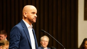 Hombre calvo con barba y traje azul hablando en un micrófono. Se ven personas borrosas en el fondo de un auditorio oscuro