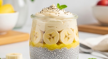 Vaso de vidrio con capas de natilla de semillas de chía, plátano en rodajas, puré amarillo, crema, semillas de chía y hojas de menta.