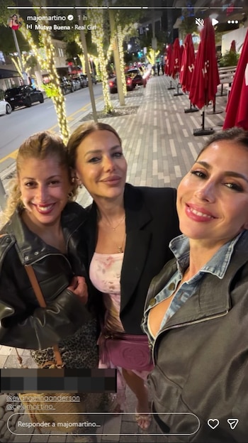 Tres mujeres sonrientes posan para una selfie al aire libre de noche, con árboles decorados con luces y sombrillas rojas de restaurante al fondo