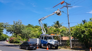 Un camión con brazo articulado eleva a un técnico hasta un poste de electricidad junto a una carretera rodeada de vegetación.